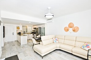 Uma sala de estar moderna com um sofá seccional bege, uma área de jantar e uma cozinha ao fundo.