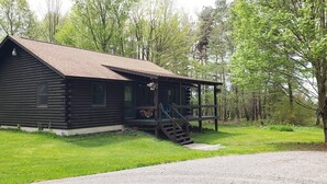 A log cabin with a porch and a staircase leading up to it, surrounded by a grassy area and trees.
