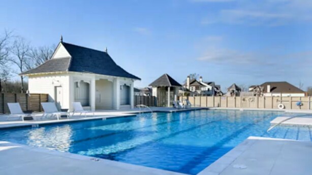 A swimming pool with lounge chairs, a small pavilion, and a fenced area with houses in the background.