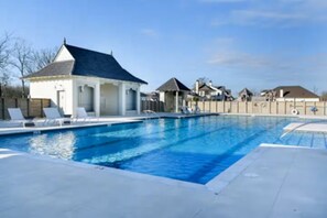 Una piscina con sillas de descanso, un pequeño pabellón y un área cercada con casas al fondo.