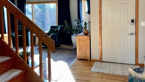 A spacious living room with a wooden staircase, a large window with curtains, a door with a handle, and a blue rug on the floor. - Welcoming retreat close to Cornell, Ithaca College, and Cayuga Lake (Ithaca)