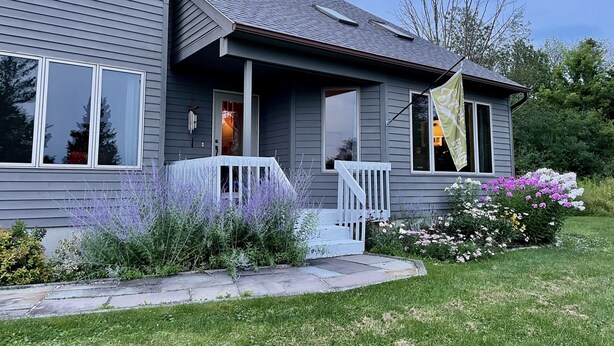 A two-story house with a grey exterior, a front porch with a white railing, and a small garden with purple flowers and a variety of other plants. - Welcoming retreat close to Cornell, Ithaca College, and Cayuga Lake (Ithaca)