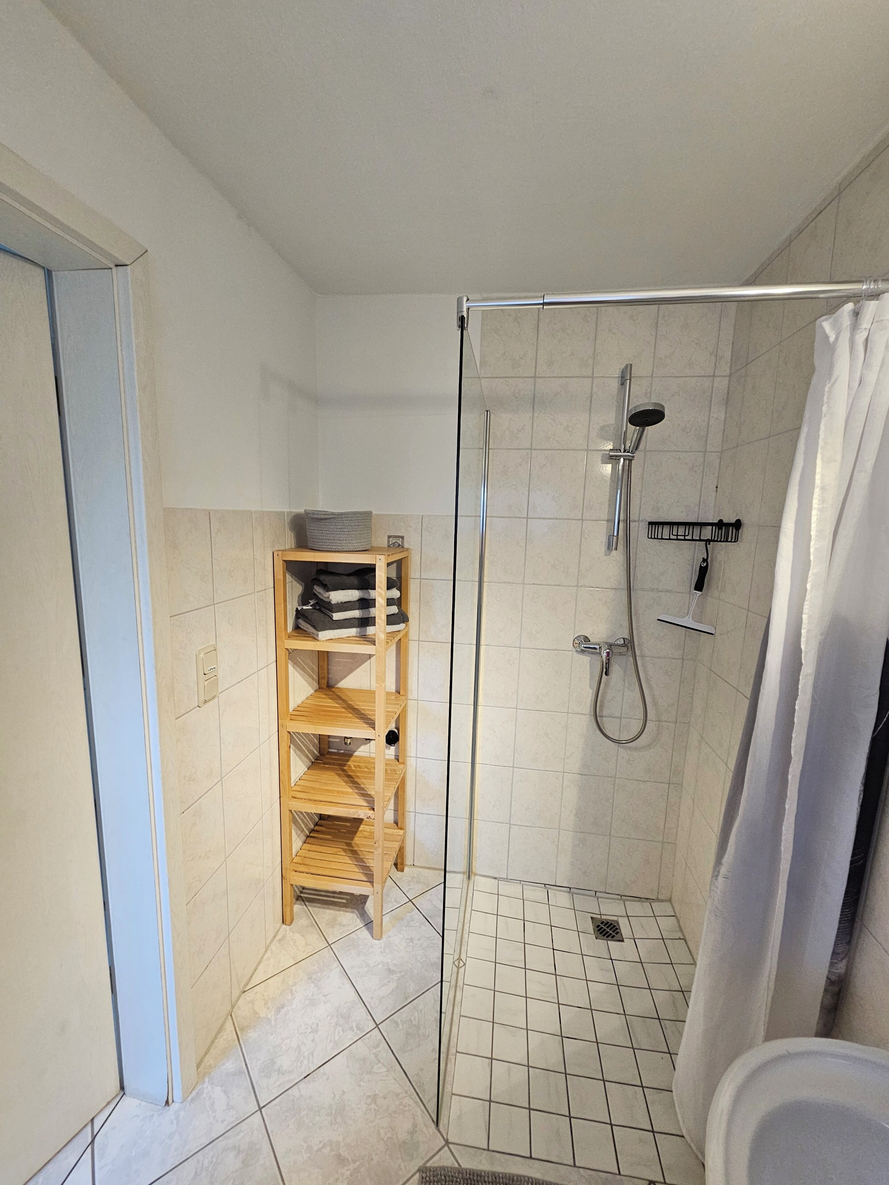 Un box de ducha con puerta de vidrio, una estantería de madera y un cabezal de ducha.