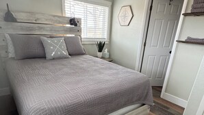 A bedroom with a bed, a nightstand, a window with blinds, a door, and a wall-mounted air conditioner. - Mil Suite-1-bedroom cottage with surrounding mountain views (La Veta)