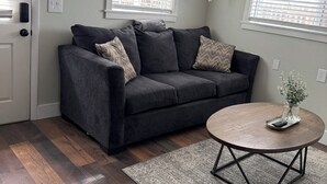 A living room with a dark grey sofa, two windows with blinds, a round coffee table with a vase, and a patterned rug. - Mil Suite-1-bedroom cottage with surrounding mountain views (La Veta)