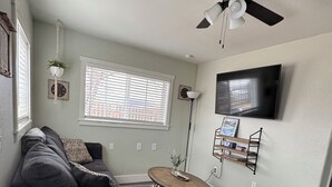 A living room with a dark grey sofa, a round coffee table, a ceiling fan, a TV mounted on the wall, and a window with blinds. - Mil Suite-1-bedroom cottage with surrounding mountain views (La Veta)