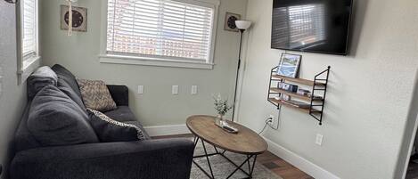 Un salon avec un canapé gris foncé, une table basse ronde, un ventilateur de plafond, un téléviseur fixé au mur et une fenêtre avec des stores.