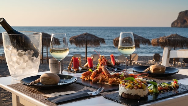 A table set with seafood, wine glasses, and a bottle of wine on a beach with umbrellas and the ocean in the background. - ISLAND CONCEPT CHANIA (Chania)
