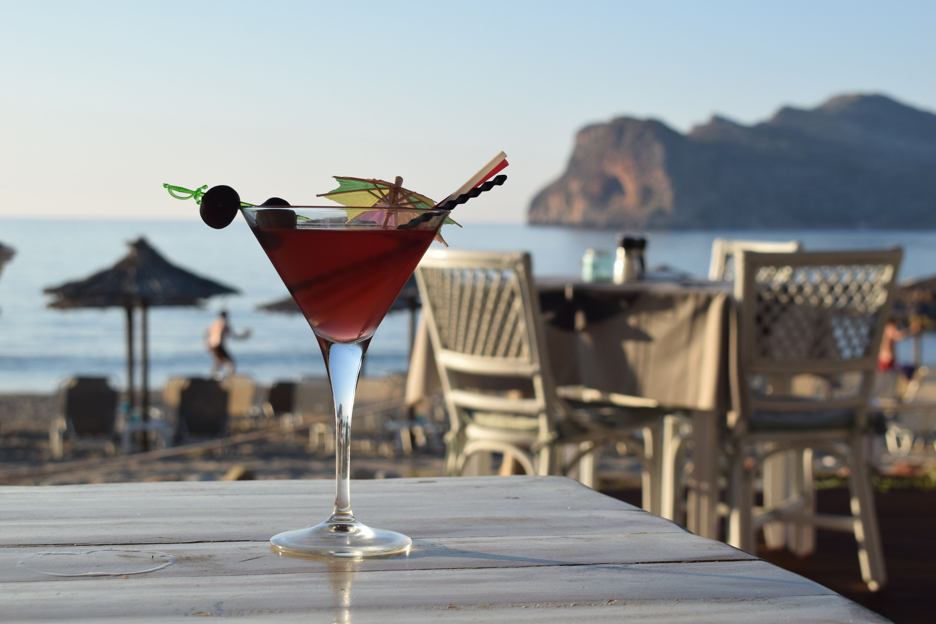 Taça de martini com enfeite sobre uma mesa à beira-mar, com cadeiras e guarda-sóis de praia ao fundo.