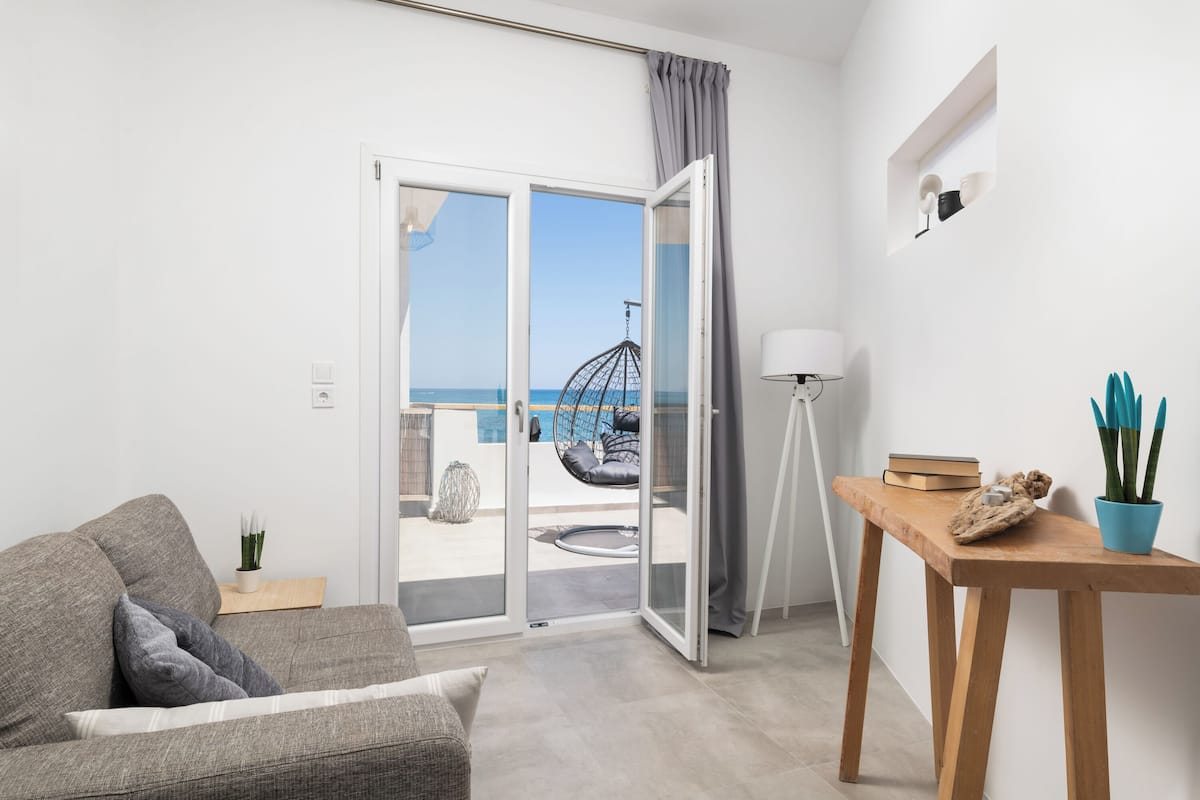 A modern living room with a grey sofa, a wooden side table, and a view of the sea through a sliding glass door.
