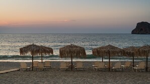 A beach with lounge chairs and straw umbrellas, a rocky outcrop in the background, and calm sea waves. - ISLAND CONCEPT CHANIA (Chania)