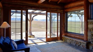 A log cabin interior with a stone fireplace, a blue sofa, and large windows offering a view of the mountains. - Glass Cottage Overlooking Beartooth Mountain Range (Roberts)