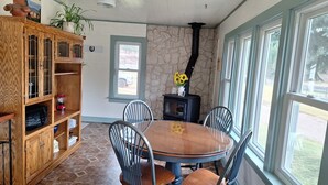 Un comedor con una mesa redonda de madera, cuatro sillas, un aparador de madera con puertas de cristal, una pared de piedra, un estufa de leña negra y una ventana con vistas al exterior.