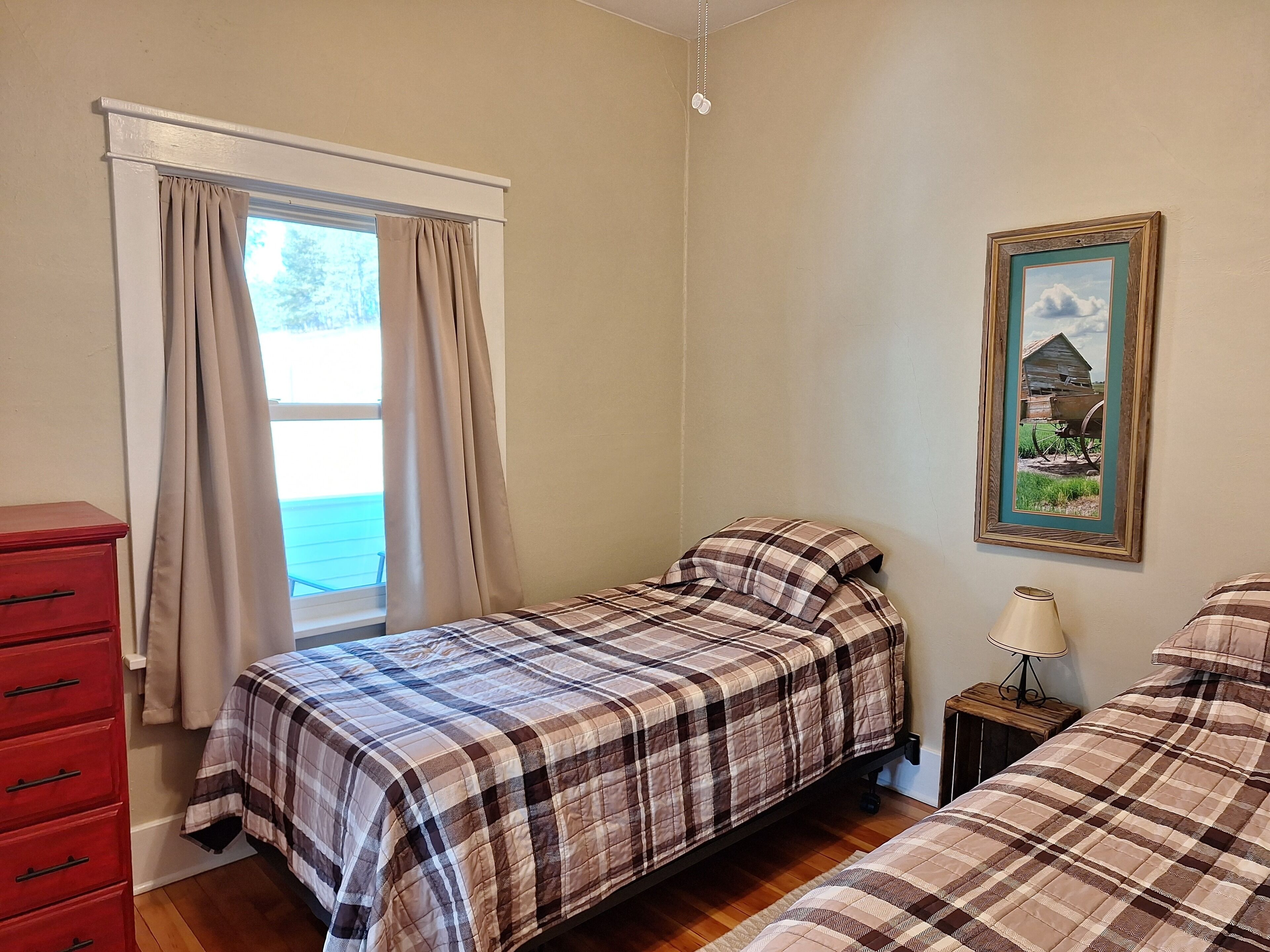 Une chambre avec deux lits, une commode, une fenêtre avec des rideaux, une lampe et un tableau encadré au mur.