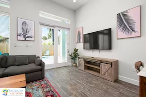 A modern living room with a grey sofa, a flat-screen TV mounted on the wall, and a wooden entertainment unit. - Coastal Getaway for 12 | Private Pool + Beach Access (South Padre Island)
