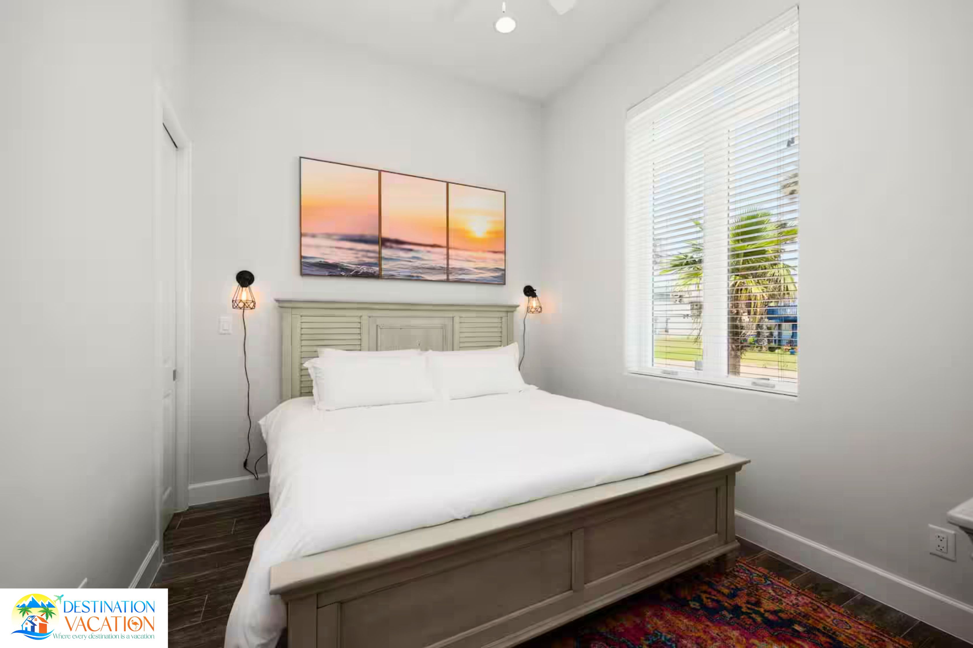 A bedroom with a bed, a window with blinds, a painting above the bed, and a logo in the corner.