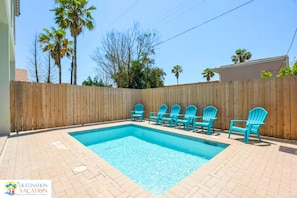 A rectangular swimming pool with a wooden fence surrounding it, and several blue chairs arranged around the pool. - Coastal Getaway for 12 | Private Pool + Beach Access (South Padre Island)