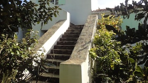 Eine Treppe führt zu einem weißen Gebäude mit grünen Fensterläden, umgeben von viel Grün.