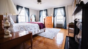 A bedroom with a wooden floor, a bed with a floral bedspread, a dresser, a mirror, and a fireplace. - The Clipper - 1747 Historic Isaac Pierce House 1st Floor (Somerset)