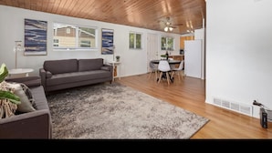 Uma sala de estar ampla com teto de madeira, um sofá cinza, um espaço de refeições com mesa e cadeiras, e uma janela grande.