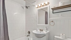 Um banheiro com azulejos brancos, uma banheira, pia com espelho e vaso sanitário.