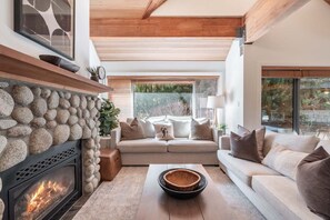 Un salon chaleureux avec une cheminée en pierre, un ensemble de canapés et une grande fenêtre donnant sur un paysage verdoyant.