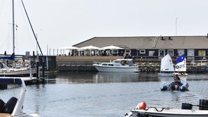 En lysthavn med både, en sejlbåd med blå og hvidt flag samt en bygning med udendørs siddepladser.