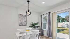 A dining area with a white table and chairs, a hanging light fixture, a potted plant, and a view through a glass door to the outside.