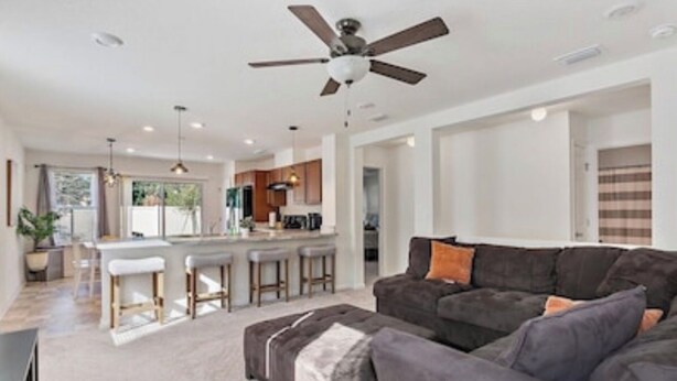 A spacious living area with a dark grey sofa, a ceiling fan, and a kitchen in the background.