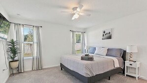 A bedroom with a bed, a ceiling fan, a nightstand, a lamp, a potted plant, and a window with curtains.