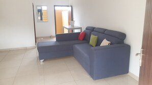 Ein modernes Wohnzimmer mit einem blauen Ecksofa, einem kleinen Tisch mit Topfpflanze und einer Tür zu einem anderen Raum.