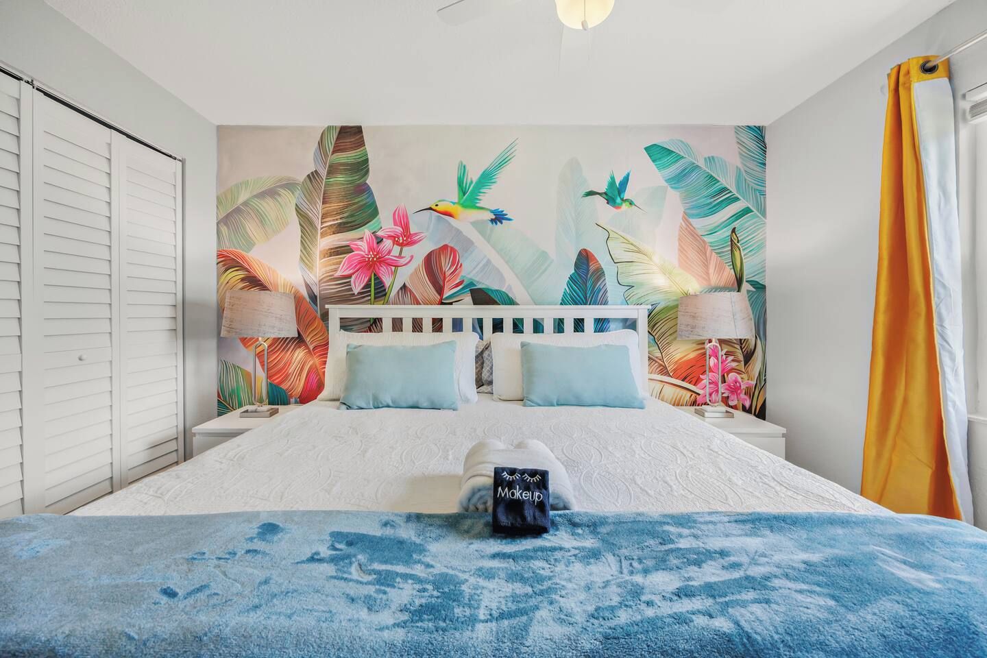 Ein Schlafzimmer mit einem Bett, Kissen, einem Nachttisch, einer Lampe und einem Wandbild mit tropischem Motiv.
