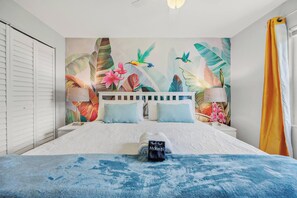 A bedroom with a bed, pillows, a nightstand, a lamp, and a tropical-themed wall mural.