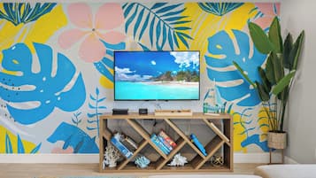 Een woonkamer met een flatscreen televisie op een houten standaard, een boekenkast met boeken en decoratieve objecten, een grote potplant en een kleurrijke, tropisch gethematiseerde muurschildering.