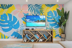 A living room with a flat-screen TV on a wooden stand, a bookshelf with books and decorative items, a large potted plant, and a colorful tropical-themed wall mural.