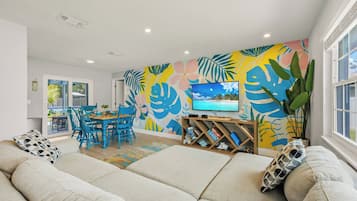 Een woonkamer met een bank, een televisie, een eethoek met stoelen en een kleurrijke muurschildering.