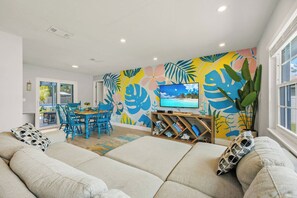 A living room with a sofa, a TV, a dining area with chairs, and a colorful wall mural.