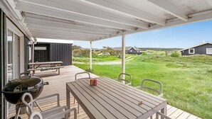 A covered outdoor area with a wooden table and chairs, a barbecue grill, and a view of a grassy field and houses in the distance. - Cozy home in Thisted with WiFi (Thisted)