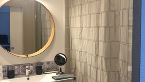 Et badeværelse med et rundt spejl, et lille rundt spejl, en vask med toiletartikler og et bruseforhæng med geometrisk mønster.