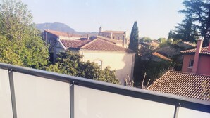 En balkon med et bord dækket til morgenmad, inklusive croissanter og appelsinjuice, med udsigt over et boligområde med huse og træer.