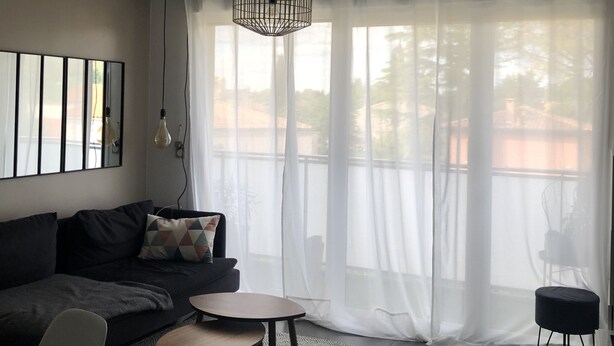 En moderne stue med en mørk sofa, et rundt sofabord og et stort vindue med lette gardiner.