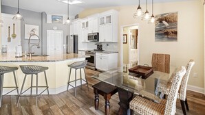 A modern kitchen with white cabinets, a central island with bar stools, and a dining area with wicker chairs. - Gray Dolphin — Waterfront Home with Heated Saltwater Pool, Dock & Kayaks (Apollo Beach)