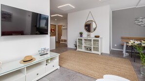 A modern living room with a flat-screen TV, a glass console table, a woven rug, and a dining area with a wooden table and chairs.