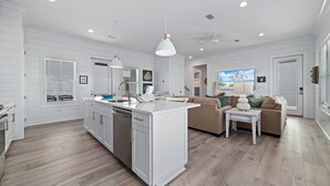 A modern kitchen with white cabinetry, a central island, and a dining area with a brown sofa. - 88° GAS Heated Pool, 85" TV, 3 King Beds, Steps to Beach, Arcade, Shuffleboard (Gulf Shores)