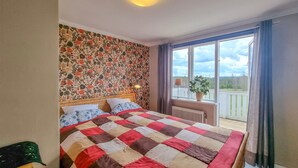 Une chambre avec un grand lit, un papier peint à motifs floraux, une vue sur l’extérieur par une fenêtre et une plante en pot sur le rebord de la fenêtre.