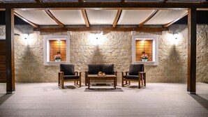 Une terrasse extérieure avec des murs en pierre, des meubles en bois et deux fenêtres ornées de rideaux.