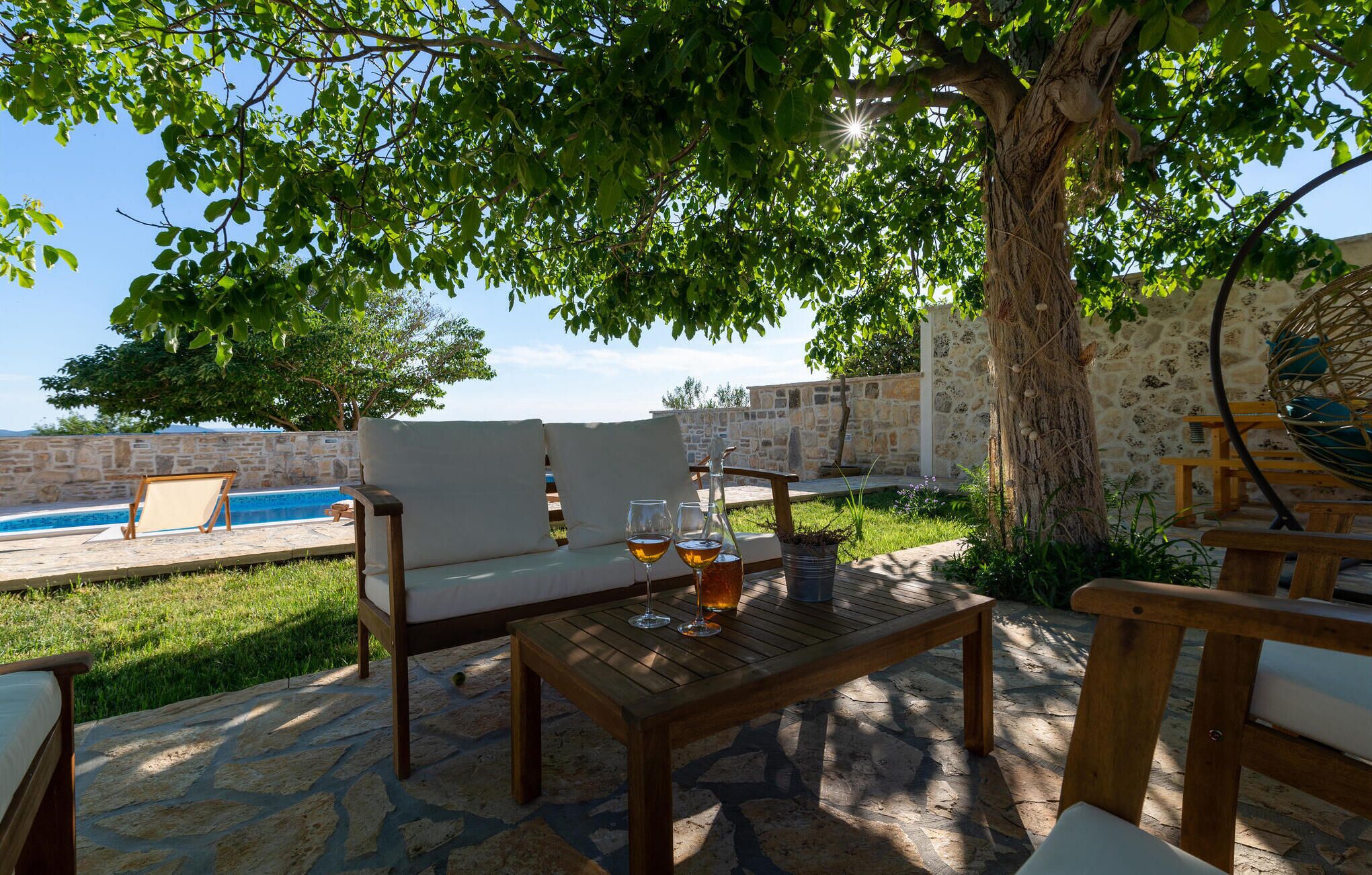 Un espacio de estar al aire libre con piscina, suelo de piedra y un árbol que da sombra.