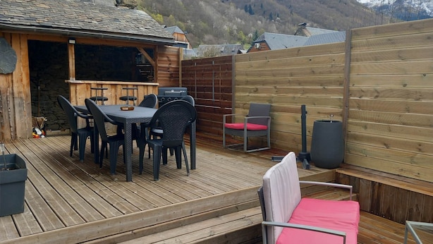 Eine Holzterrasse mit Tisch und Stühlen, einem Grill und einer Sitzecke mit Kissen.