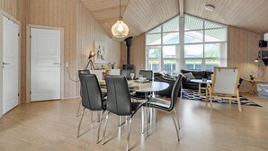 Een ruime kamer met een hoge plafond, houten wanden, een groot raam, een eettafel met stoelen, een bank en een open haard.