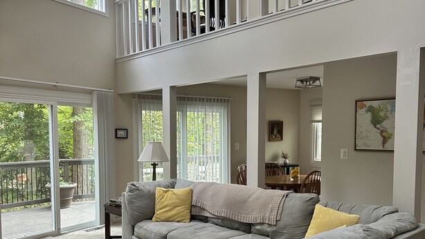 A spacious living room with a large gray sectional sofa, a ceiling fan, and a view of the outdoors through large windows. - Bay view from Calvert Cliffs, Solomons Island & State Park nearby (Lusby)